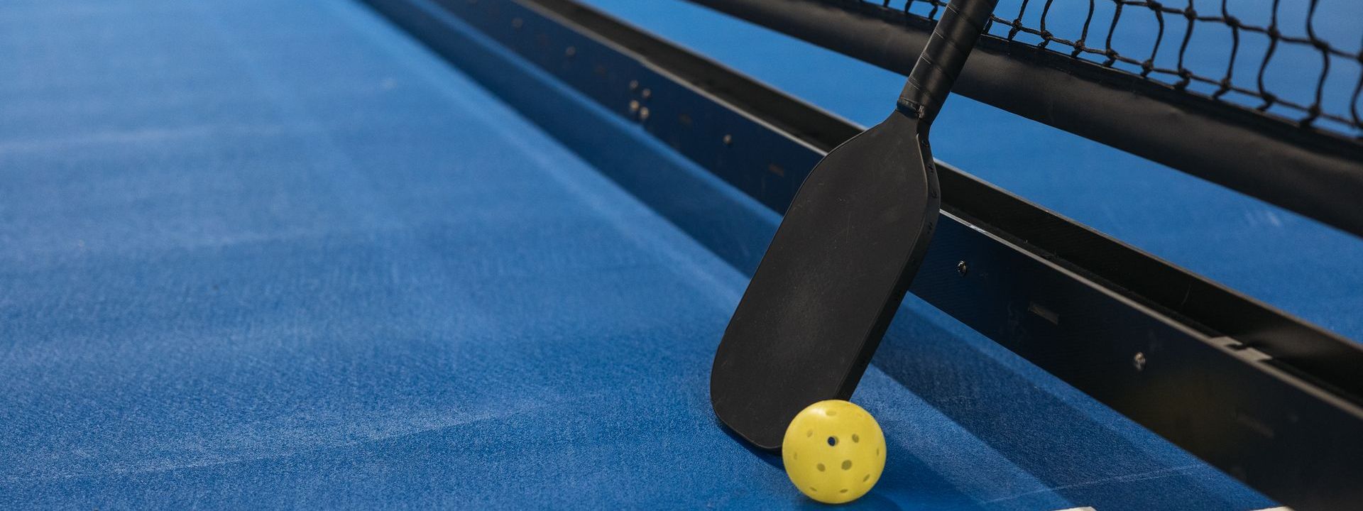 Pickleball paddle leaning against a court net with a yellow ball on a blue court